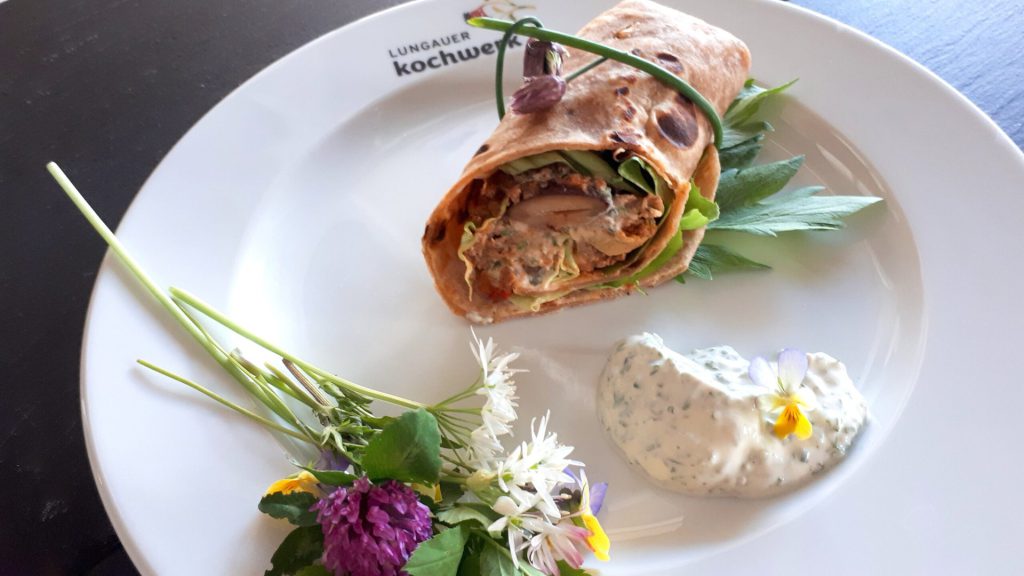 Lungau-Wrap auf einem Teller, dekoriert mit Wildkräutern und Joghurt-Dip.
